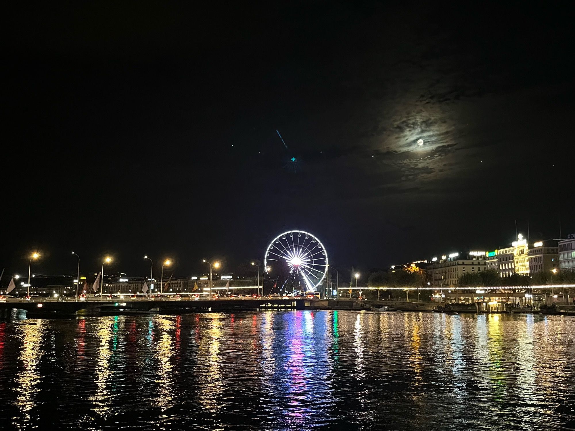 Nighttime scene of a ferris wheel illuminated by colorful lights, reflecting on the water.