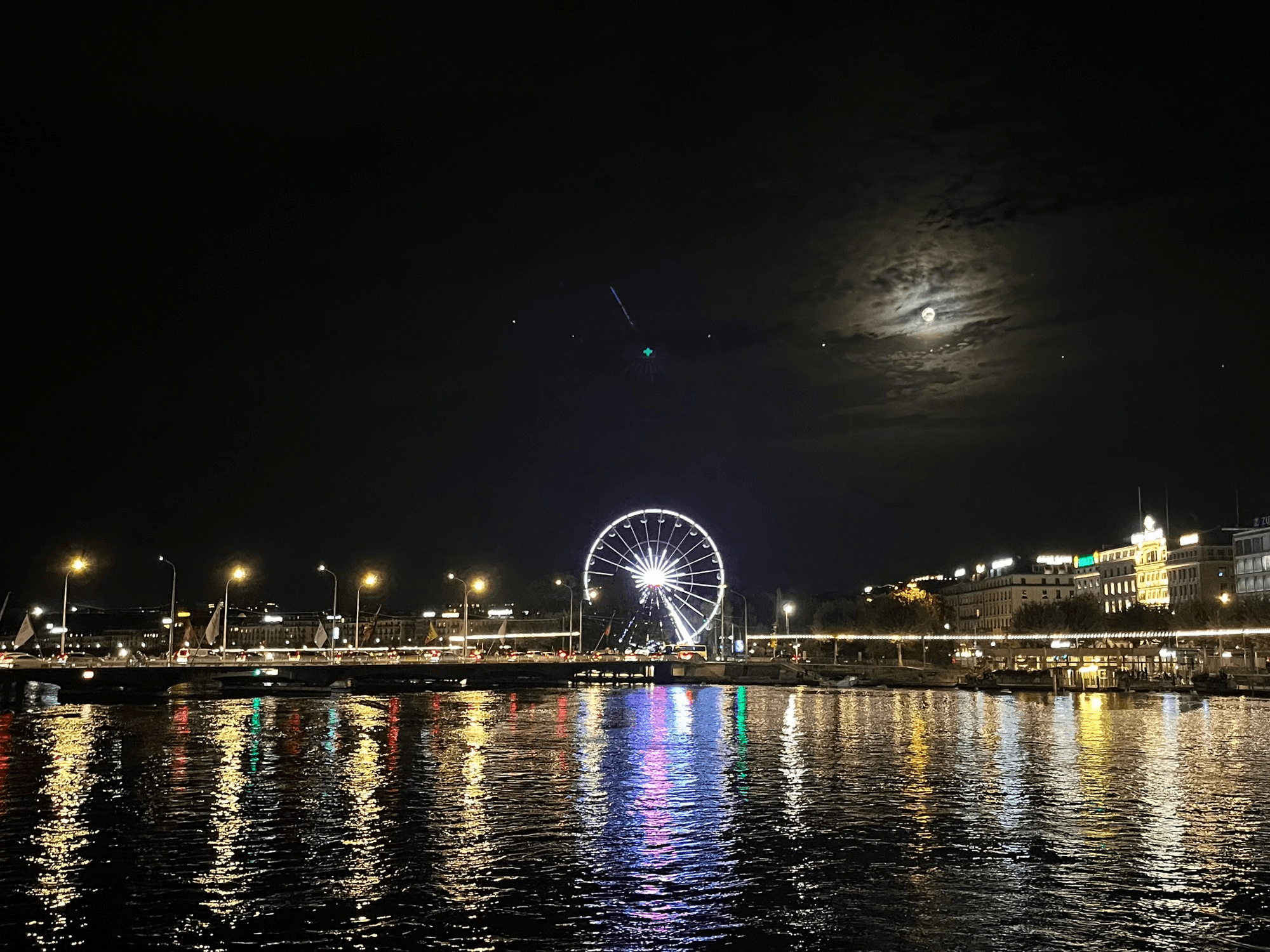 Nighttime scene of a ferris wheel illuminated by colorful lights, reflecting on the water.