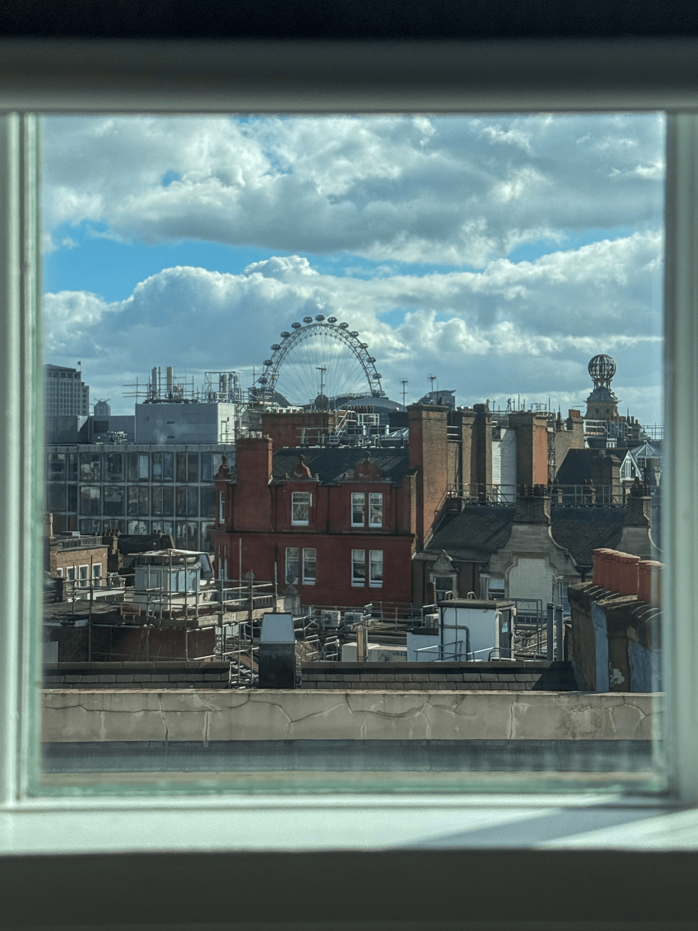 View through a window showcasing rooftops and the London Eye against a cloudy sky.