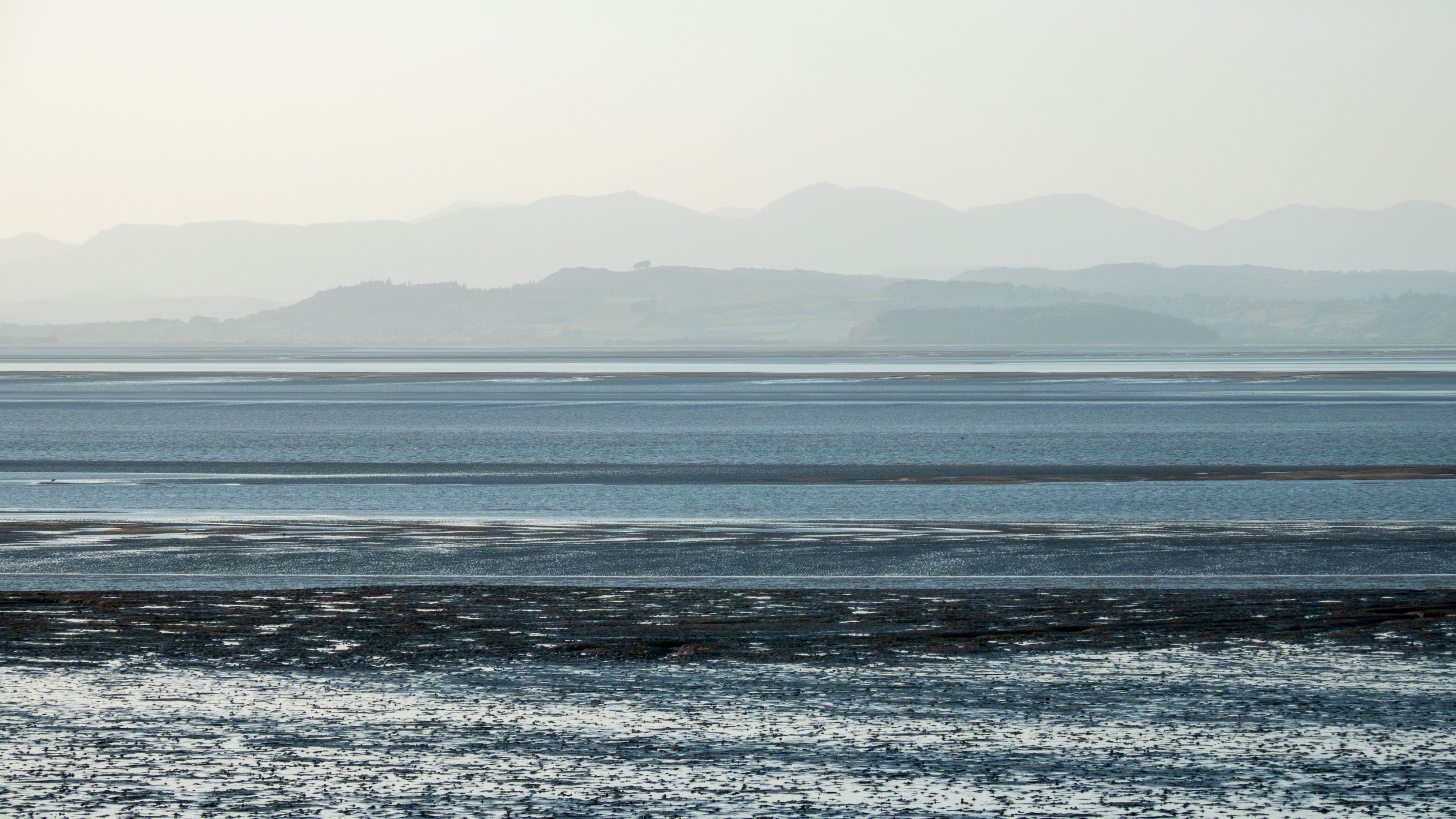 Calm seaside landscape with distant mountains and shallow water at low tide.