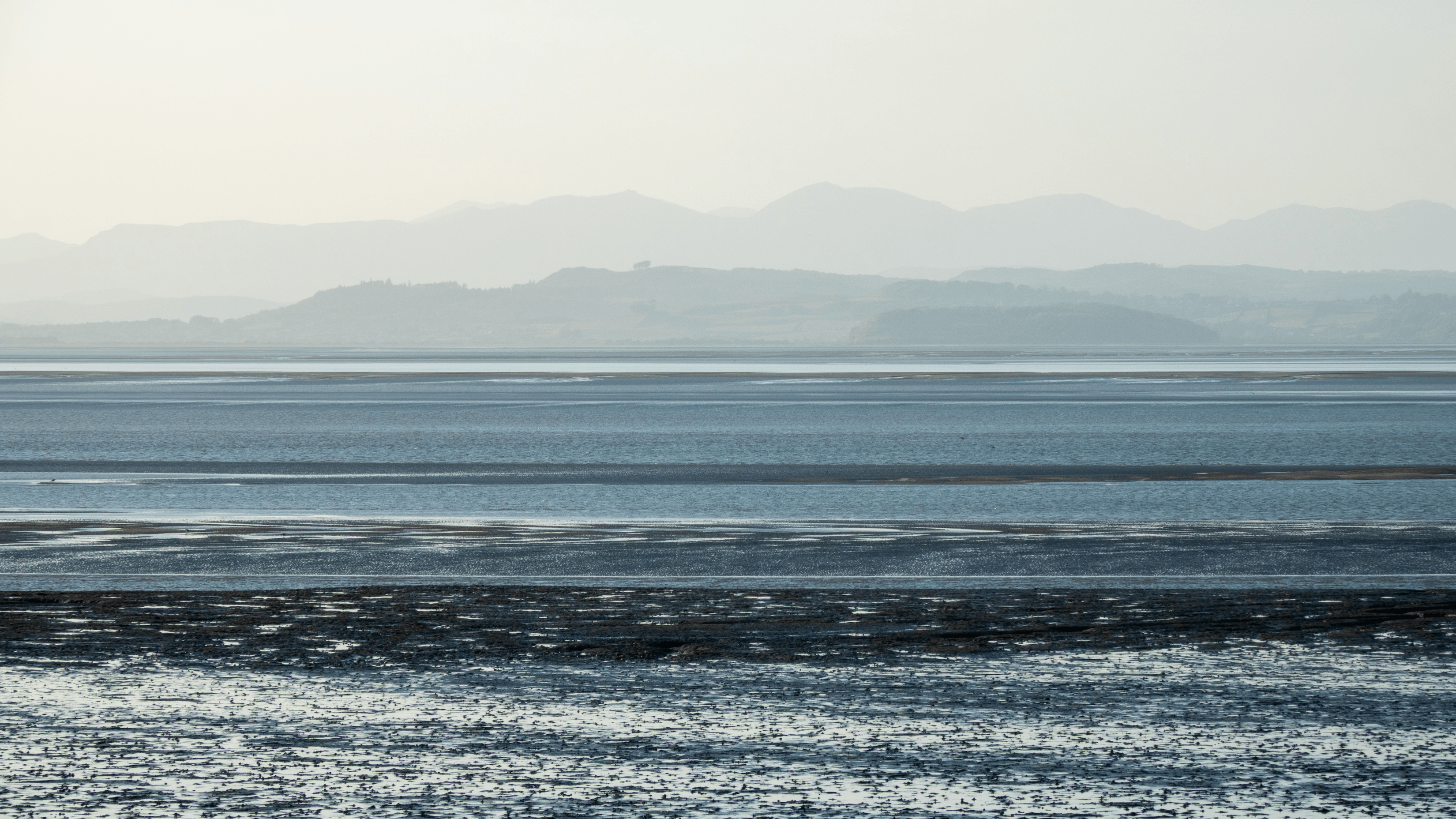 Calm seaside landscape with distant mountains and shallow water at low tide.