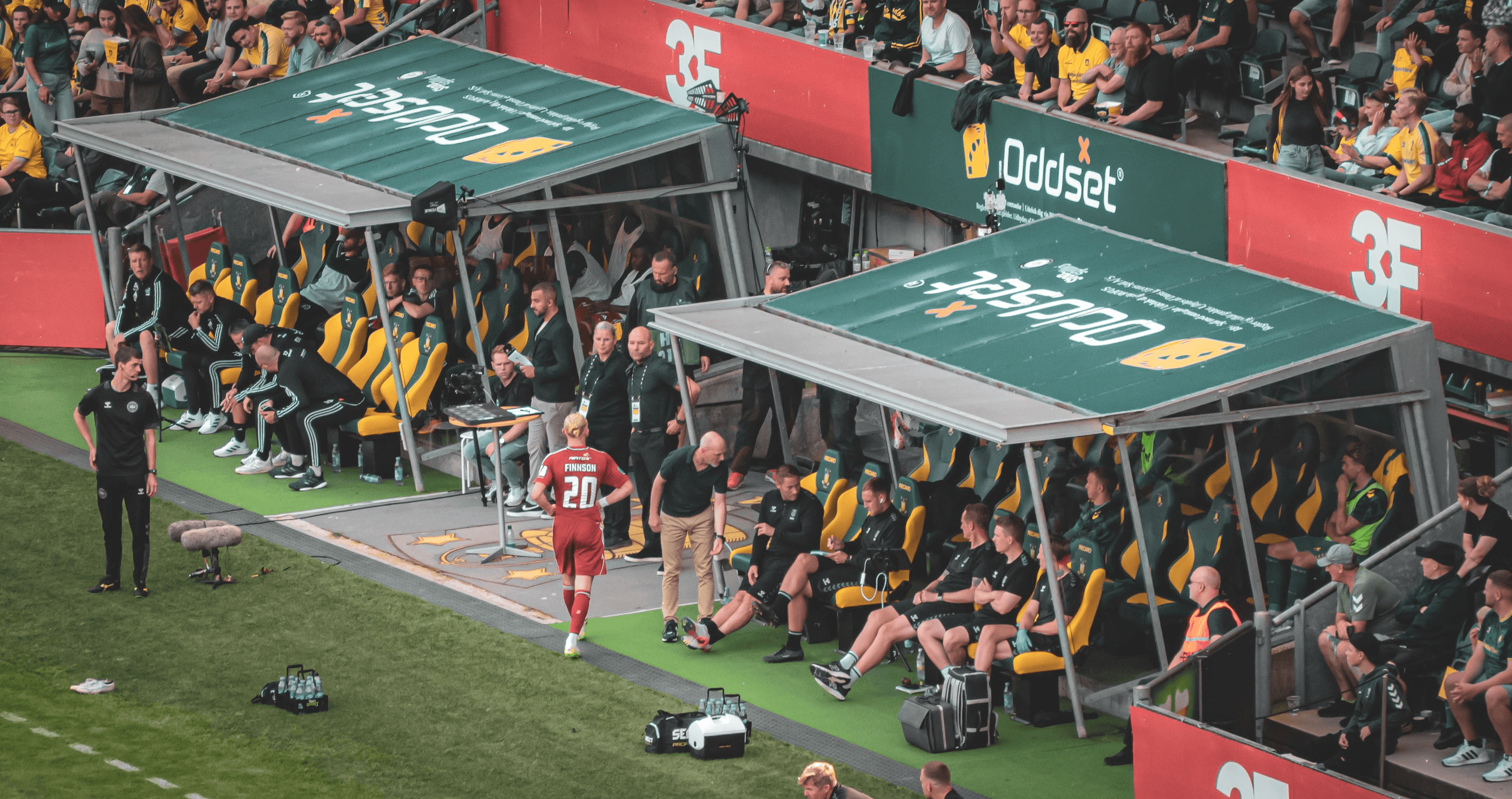 A sports bench with players sitting under canopies, while a player comes off the field.