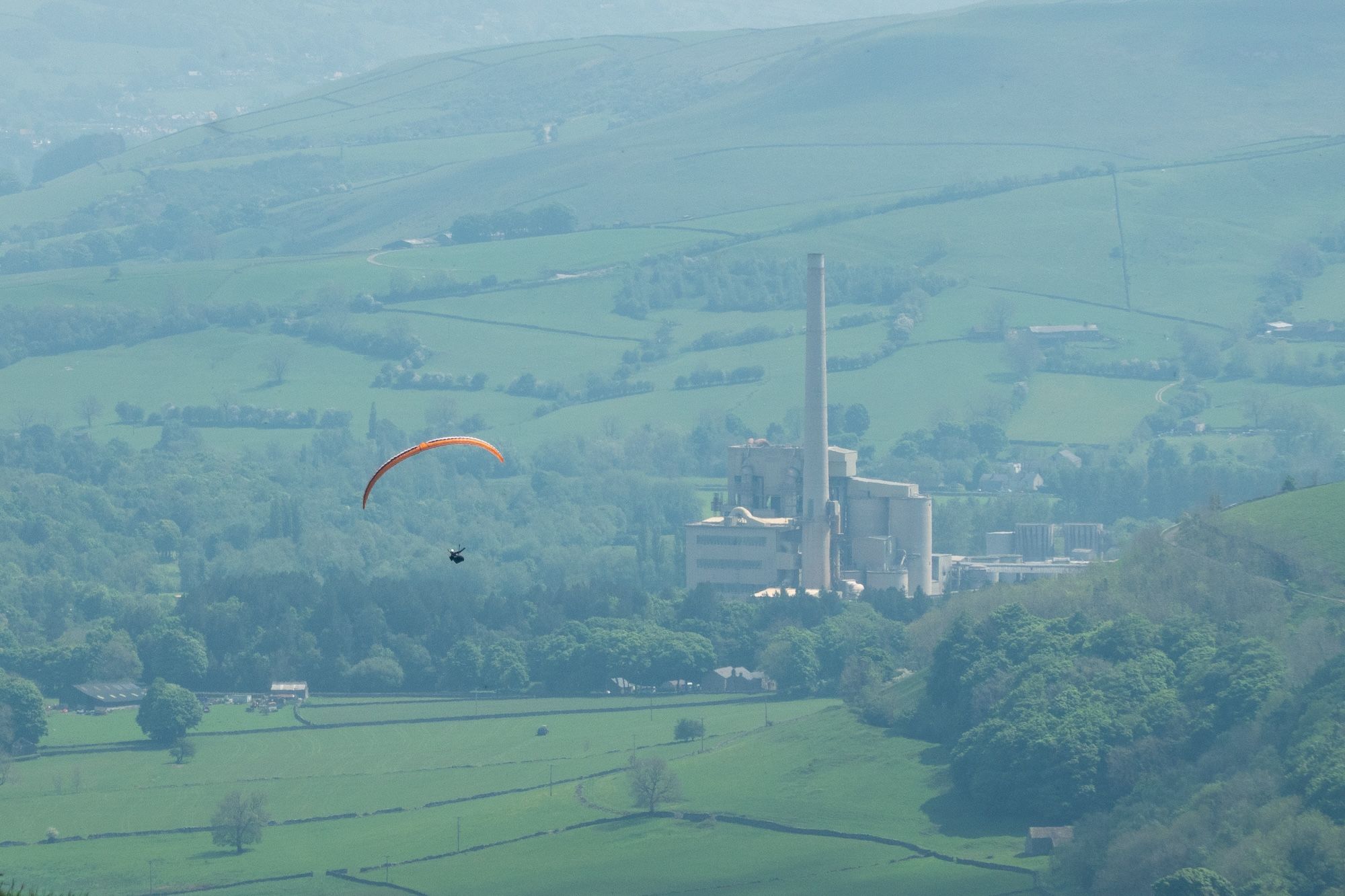 A paraglider with an orange wing soaring over green hills and a factory in the distance.