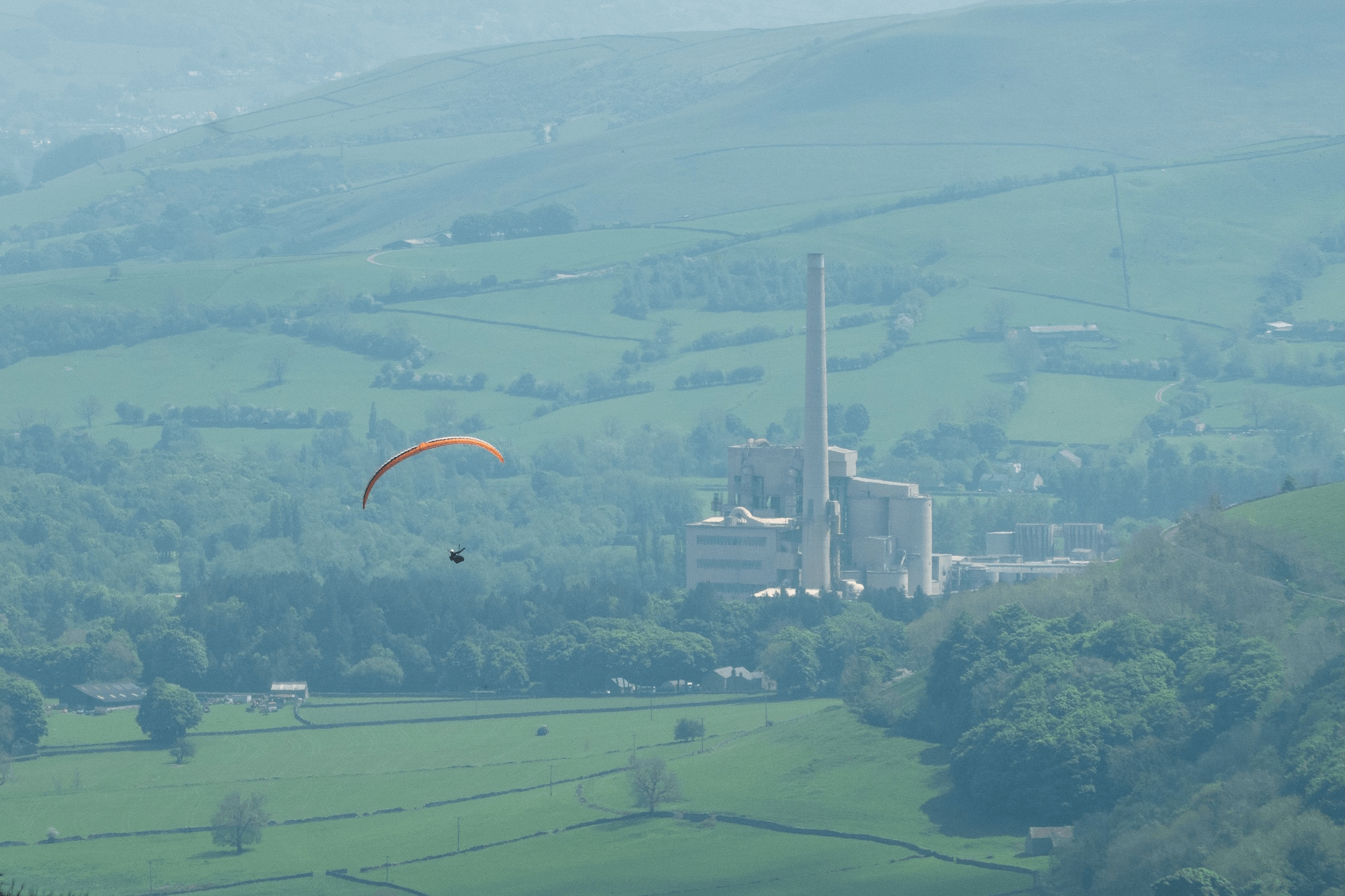 A paraglider with an orange wing soaring over green hills and a factory in the distance.